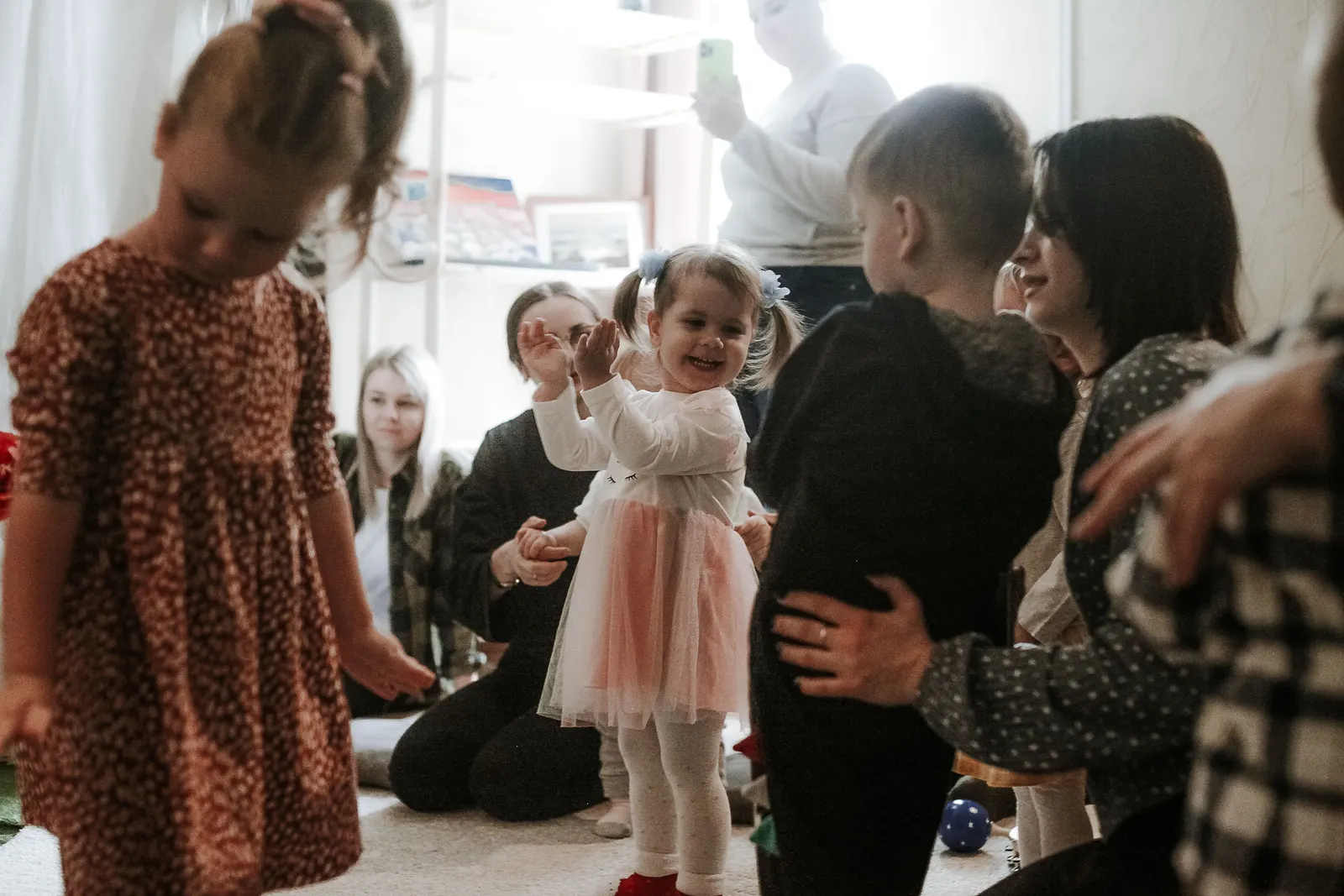 Caregivers playing with a group of smiling young children in a bright home living room